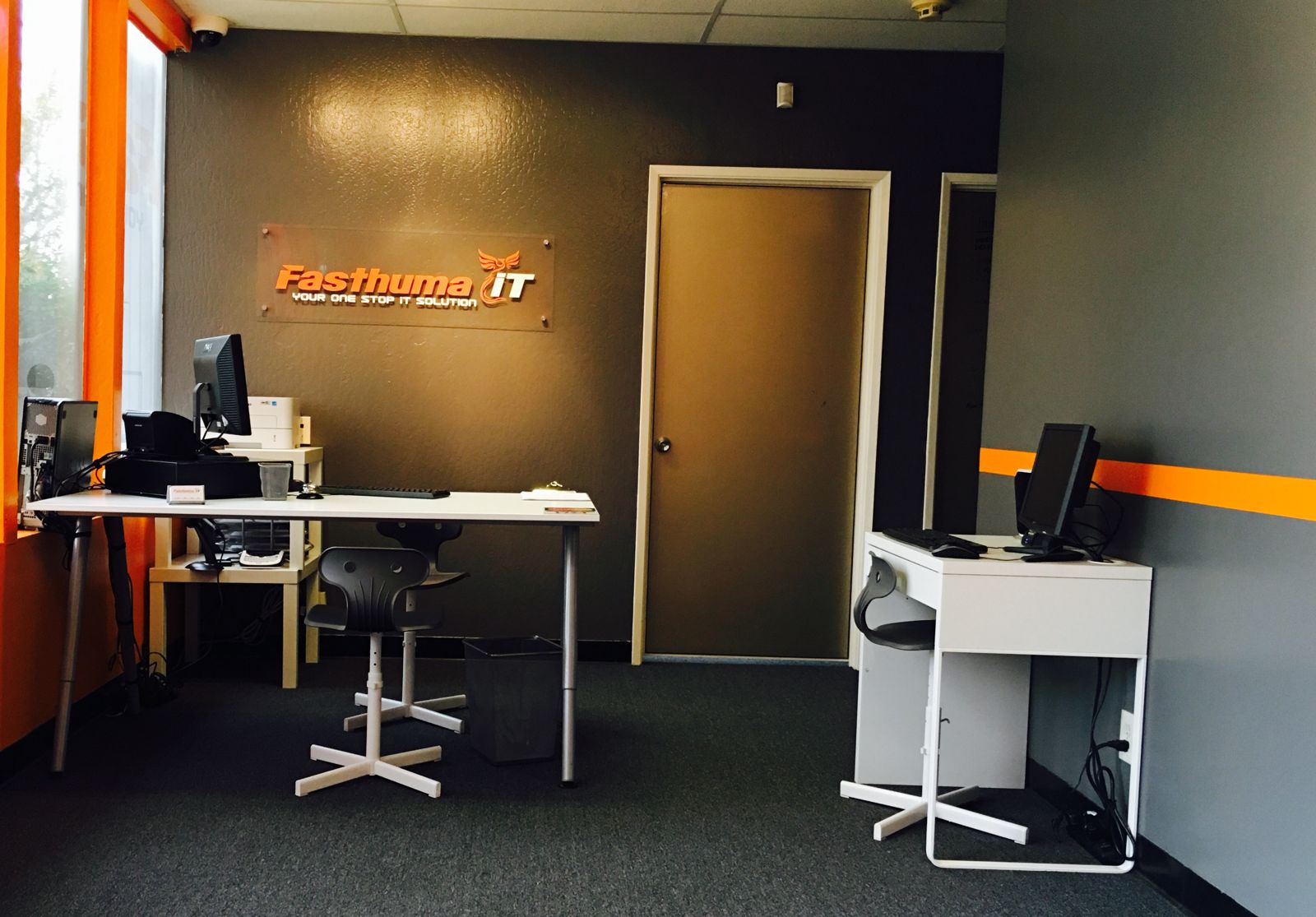 Inside the Fasthuma office at 42307 Osgood Rd, Suite B, Fremont, CA: technician desks with computers under the orange Fasthuma iT logo sign on a gray wall.